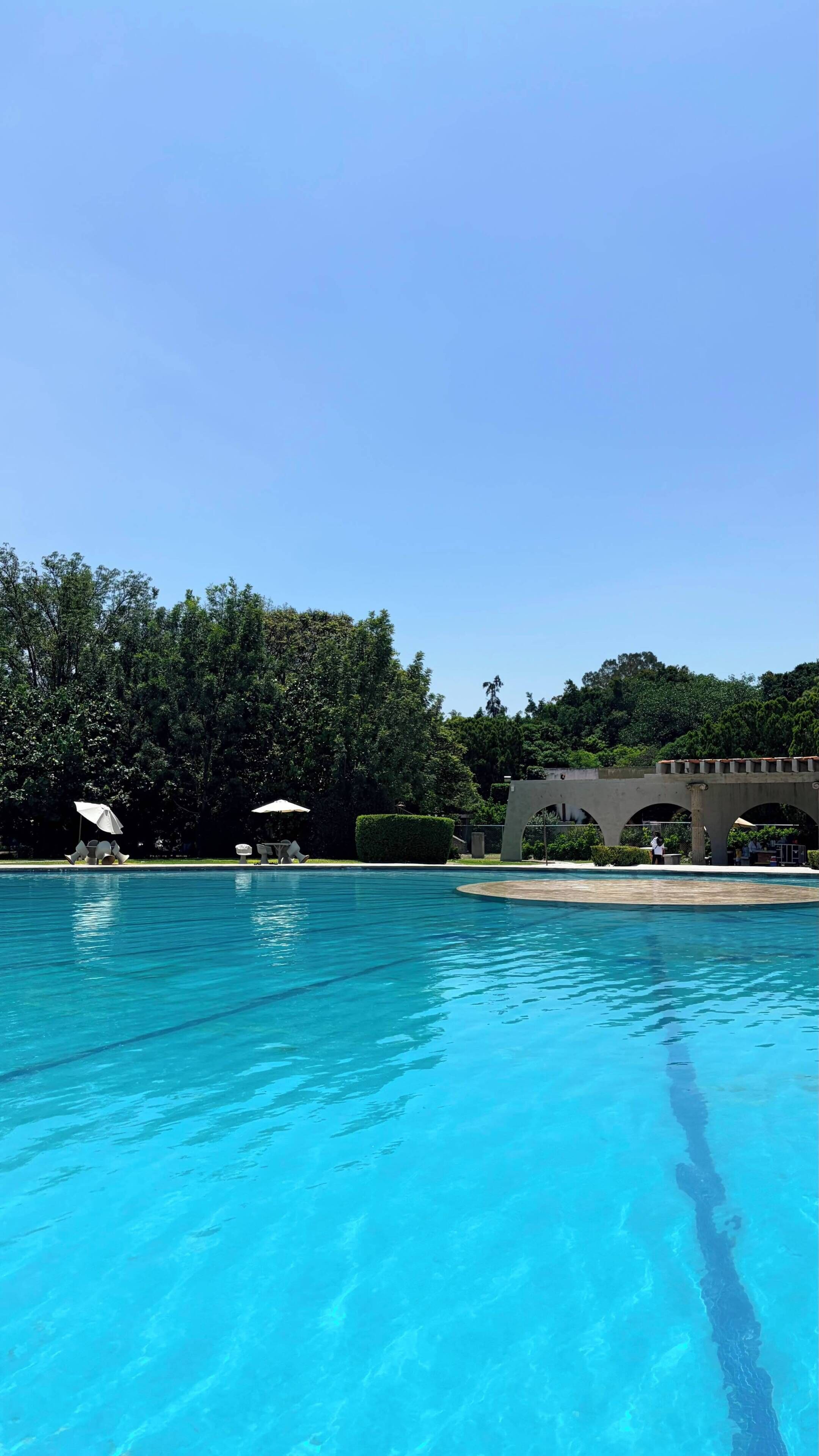 Piscina cristalina con sillas de descanso y sombrillas, rodeada de exuberante vegetación y un puente de piedra.