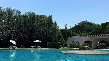 Piscina cristalina con sillas de descanso y sombrillas, rodeada de exuberante vegetación y un puente de piedra.
