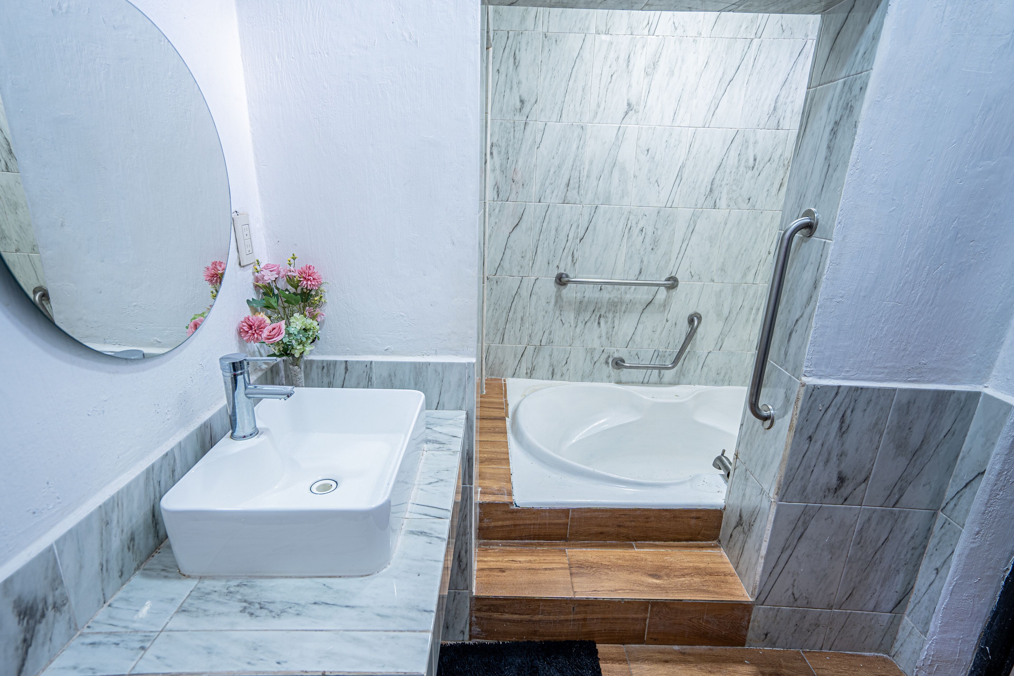 A bathroom with a white sink, a mirror, a flower vase, a bathtub, and a shower area with a handrail.