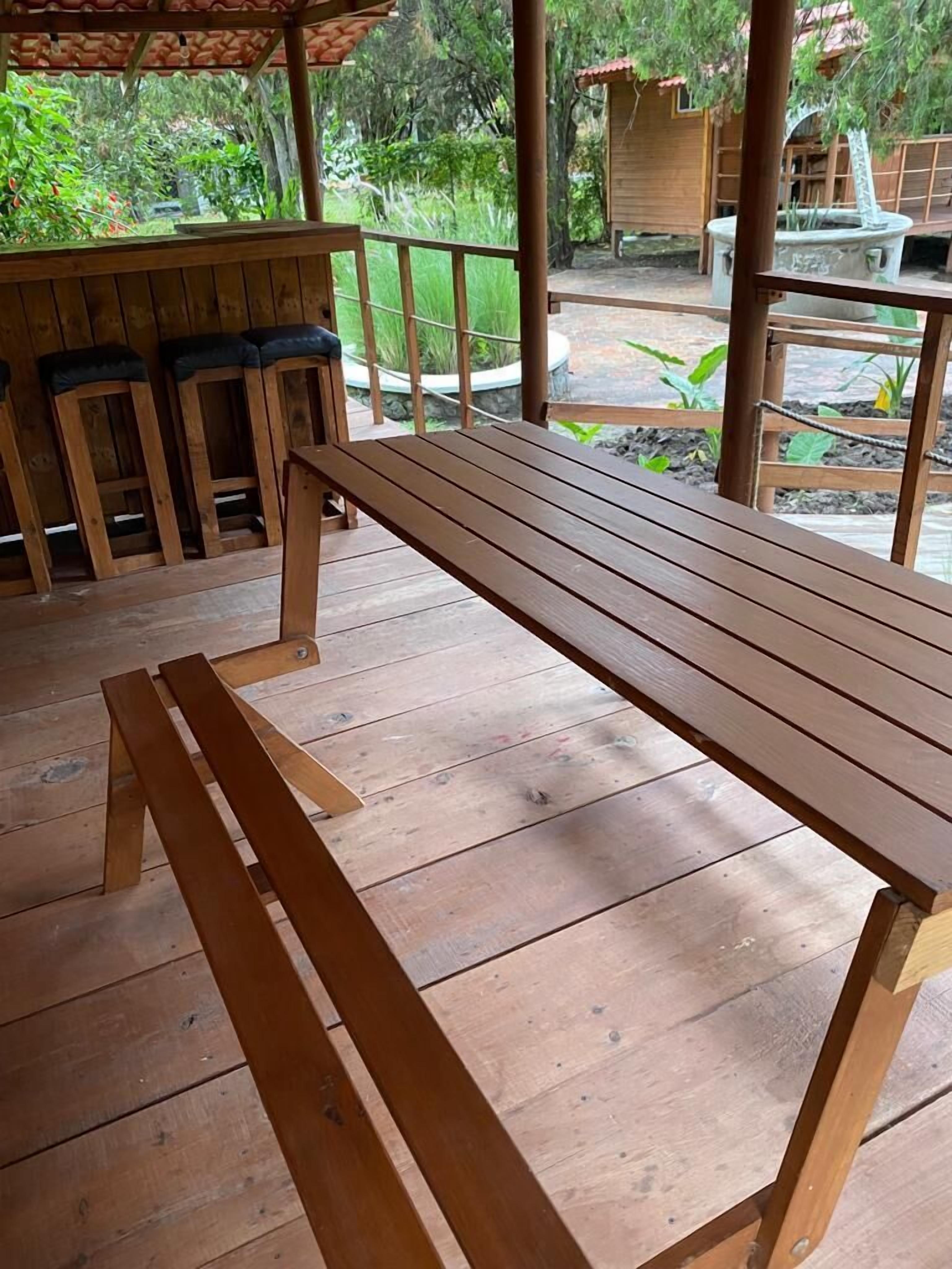 Un ensemble de table et de banc en bois est installé sur une terrasse en bois, offrant une vue sur un jardin et un bâtiment en arrière-plan.