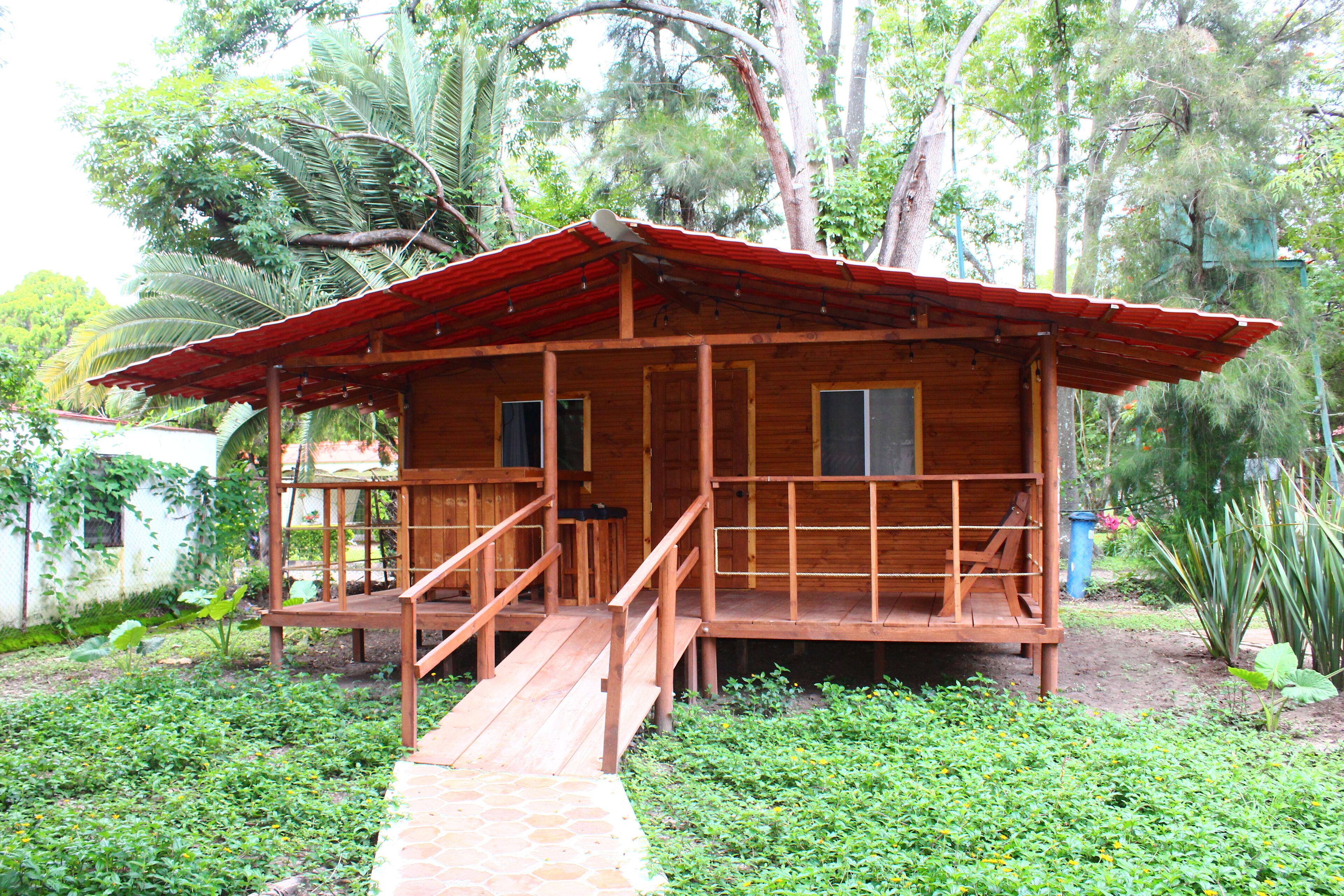 A wooden cabin with a red roof, a wooden deck, and a staircase leading to the entrance.
