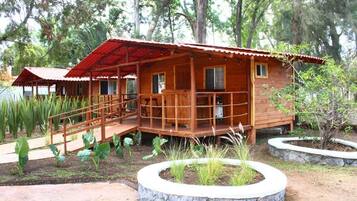 Uma cabana de madeira com varanda e um pequeno alpendre, rodeada por um jardim com várias plantas e um vaso de pedra.