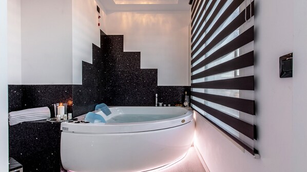A modern bathroom with a freestanding bathtub, dark tiled walls, and a large window with horizontal blinds.