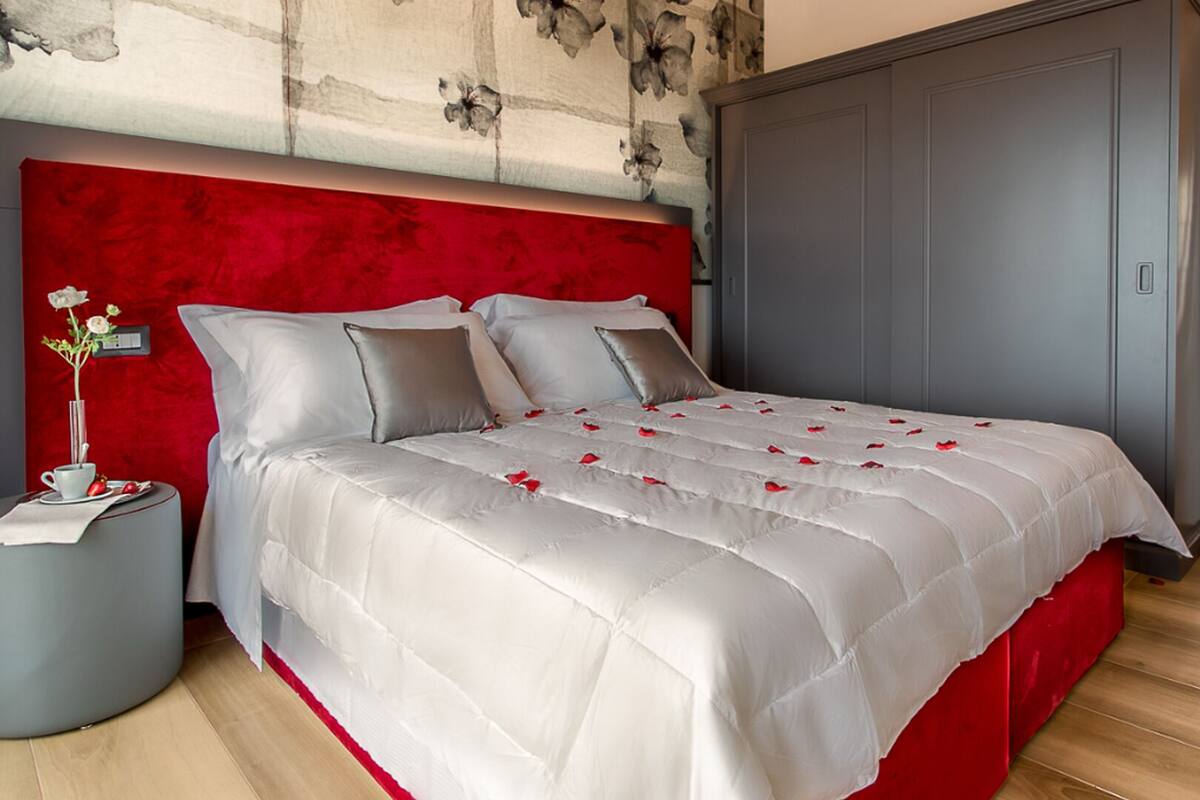 A bedroom with a large bed, a red headboard, a grey wardrobe, and a small bedside table with a vase of flowers.