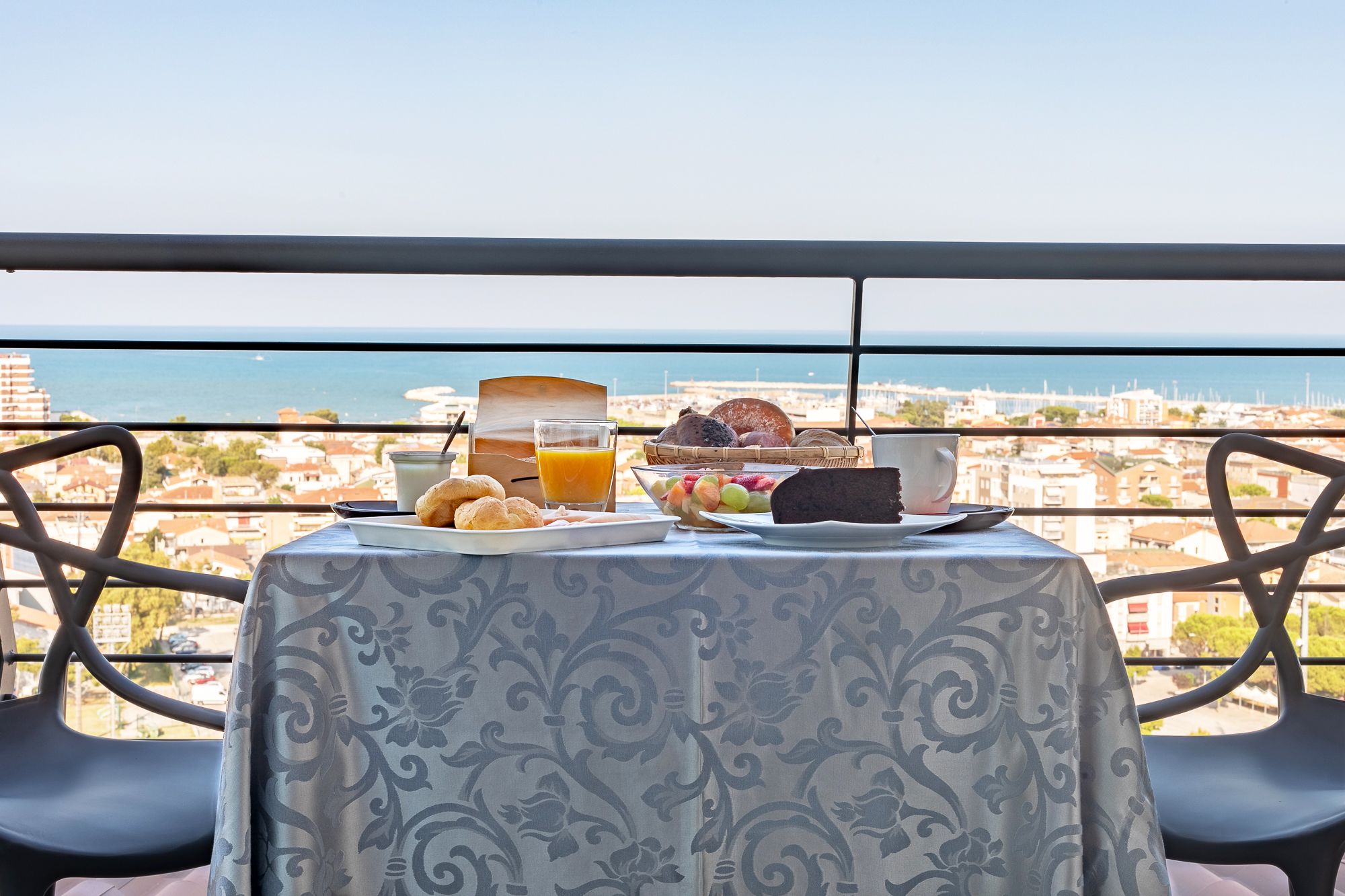 A balcony table with breakfast items, a view of the sea, and cityscape.
