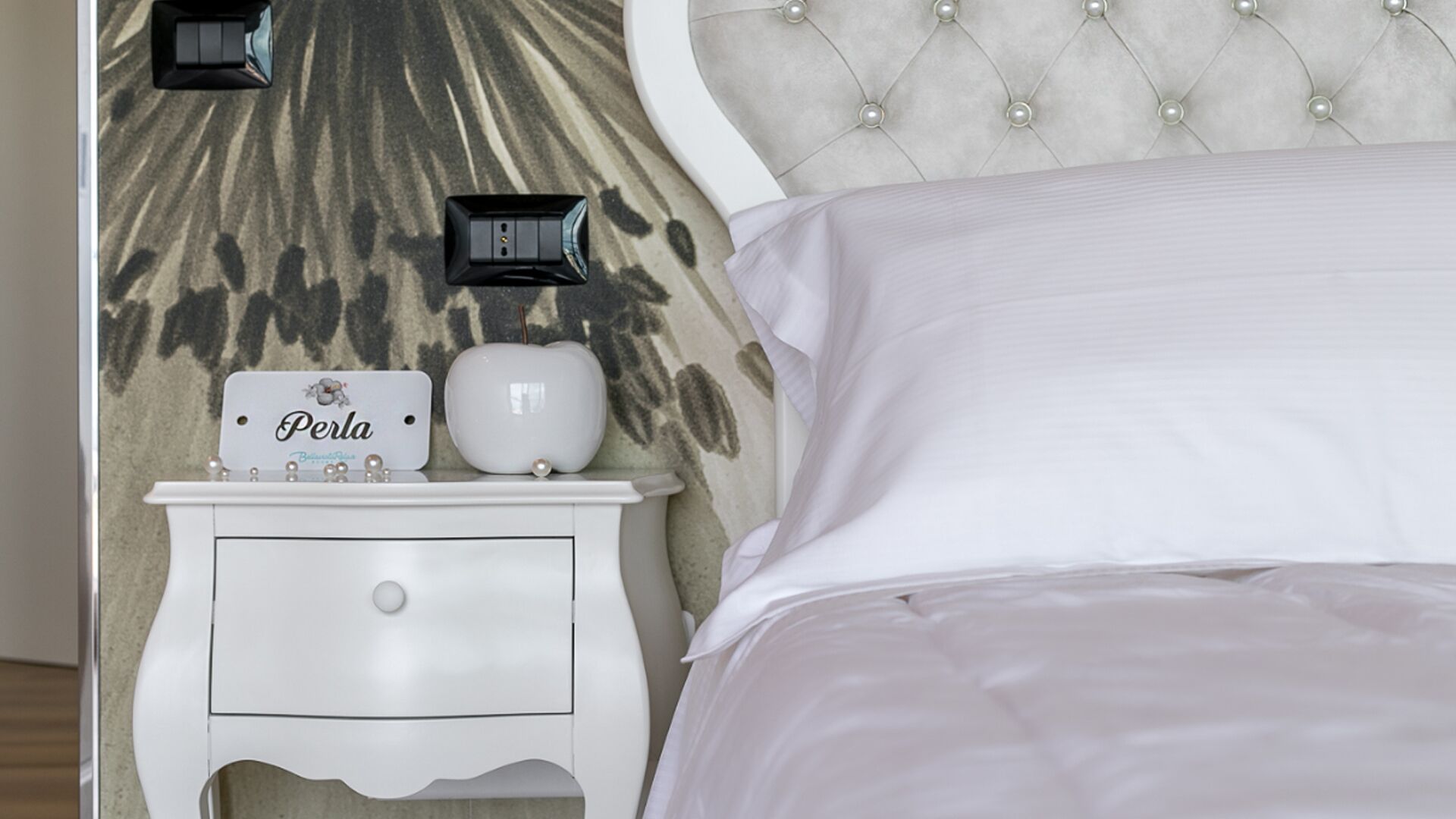 A neatly made bed with white linens, a white nightstand with a small lamp, and a wall clock.