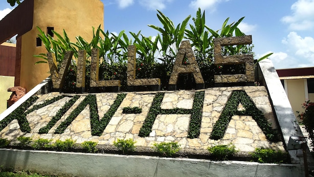 A stone wall with the words "VILLAGE KINHA" in large letters, surrounded by greenery and a clear sky. - Villas Kin Ha (Palenque)