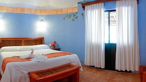 A hotel room with a large bed, a wooden bench, a small table with a vase of flowers, and a window with white curtains. - Villas Kin Ha (Palenque)