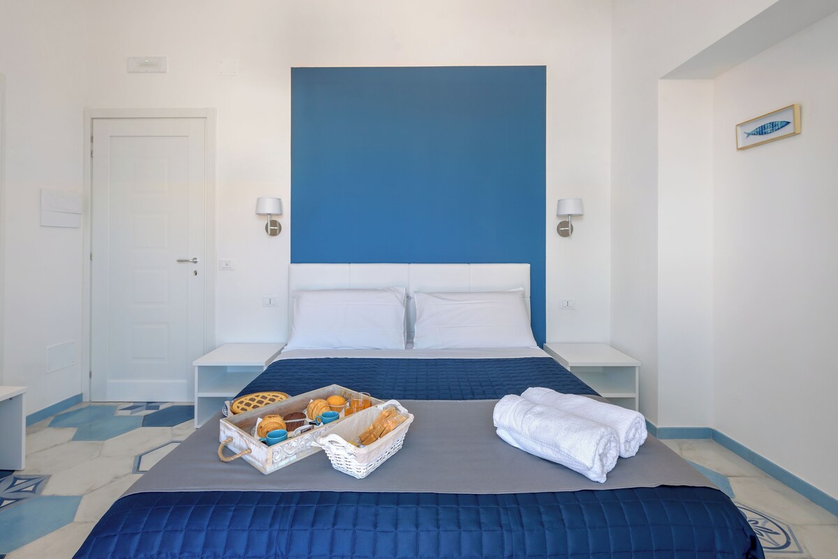 A hotel room with a large bed, a nightstand, a wall-mounted lamp, and a tray of breakfast items.