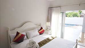 Een net opgemaakt bed met witte beddengoed en decoratieve kussens, een nachtkastje met een lamp, en uitzicht op een zwembad door een glazen deur.