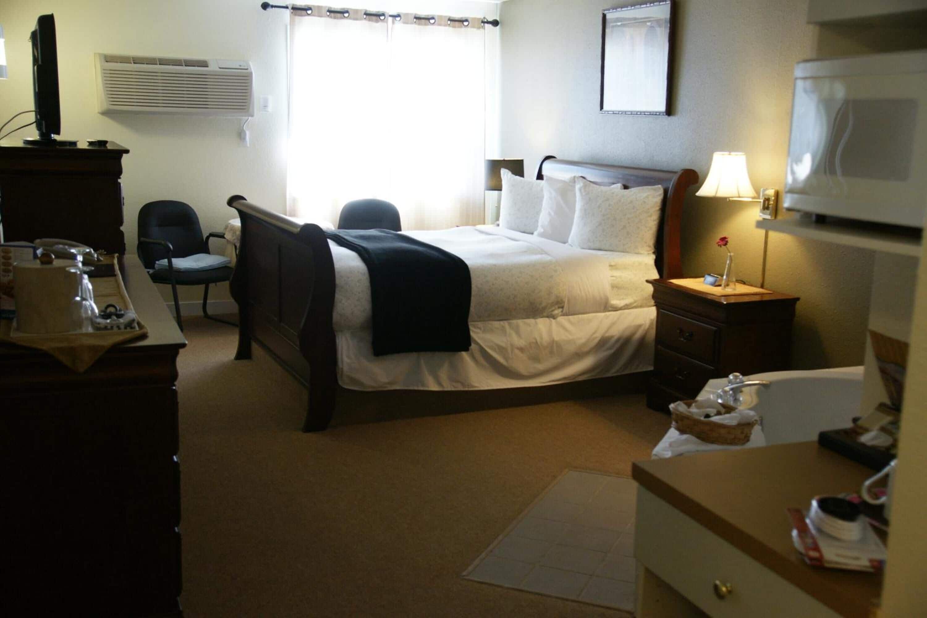 A hotel room with a bed, a desk with a computer, a chair, a bedside table with a lamp, and a television.