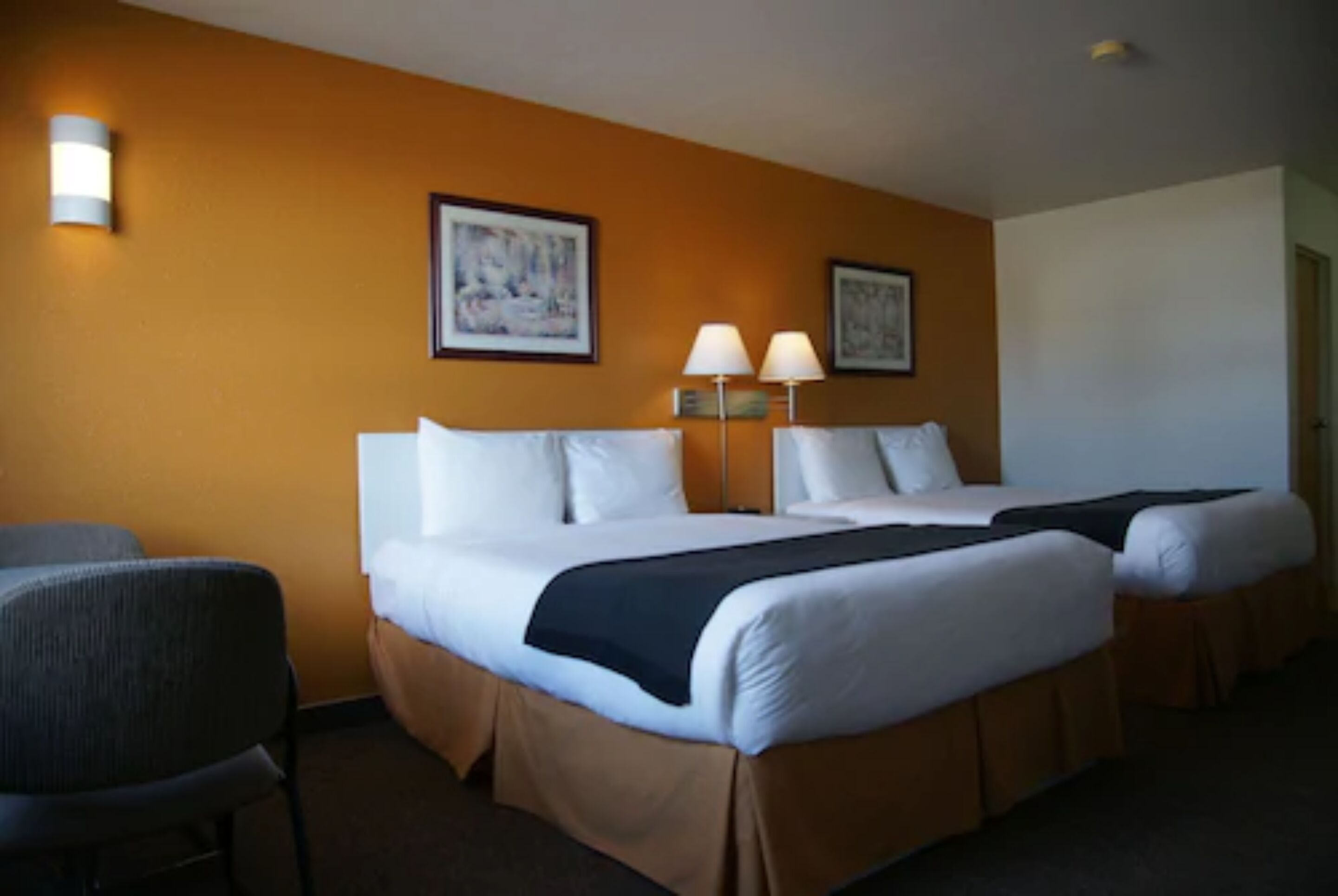 A hotel room with two beds, a chair, and two wall-mounted lamps.