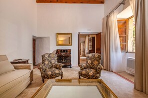 A living room with a sofa, two patterned armchairs, a glass coffee table, a wooden cabinet, and a mirror on the wall.