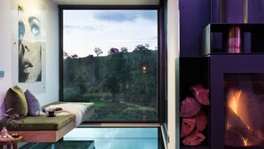 Un salon moderne doté d’un plancher en verre, d’un canapé avec des coussins, d’une cheminée et d’une grande fenêtre donnant sur un paysage naturel.