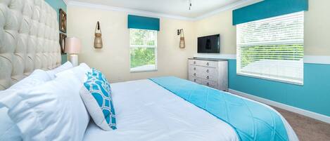 Un dormitorio con cabecera capitonada, cama con ropa de cama blanca y azul, cómoda, ventilador de techo y dos ventanas con persianas.
