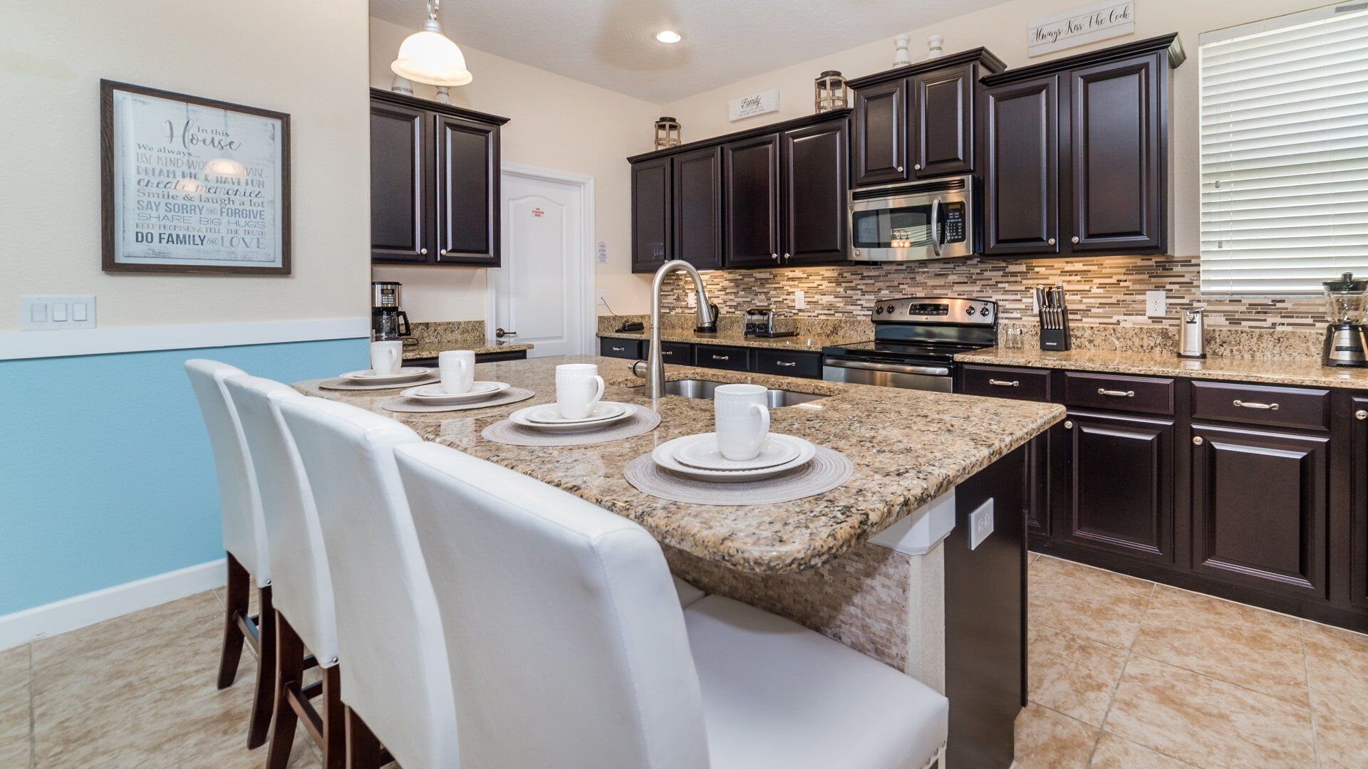 A modern kitchen with dark cabinets, granite countertops, and stainless steel appliances. There is a dining area with white chairs and a table set for two.