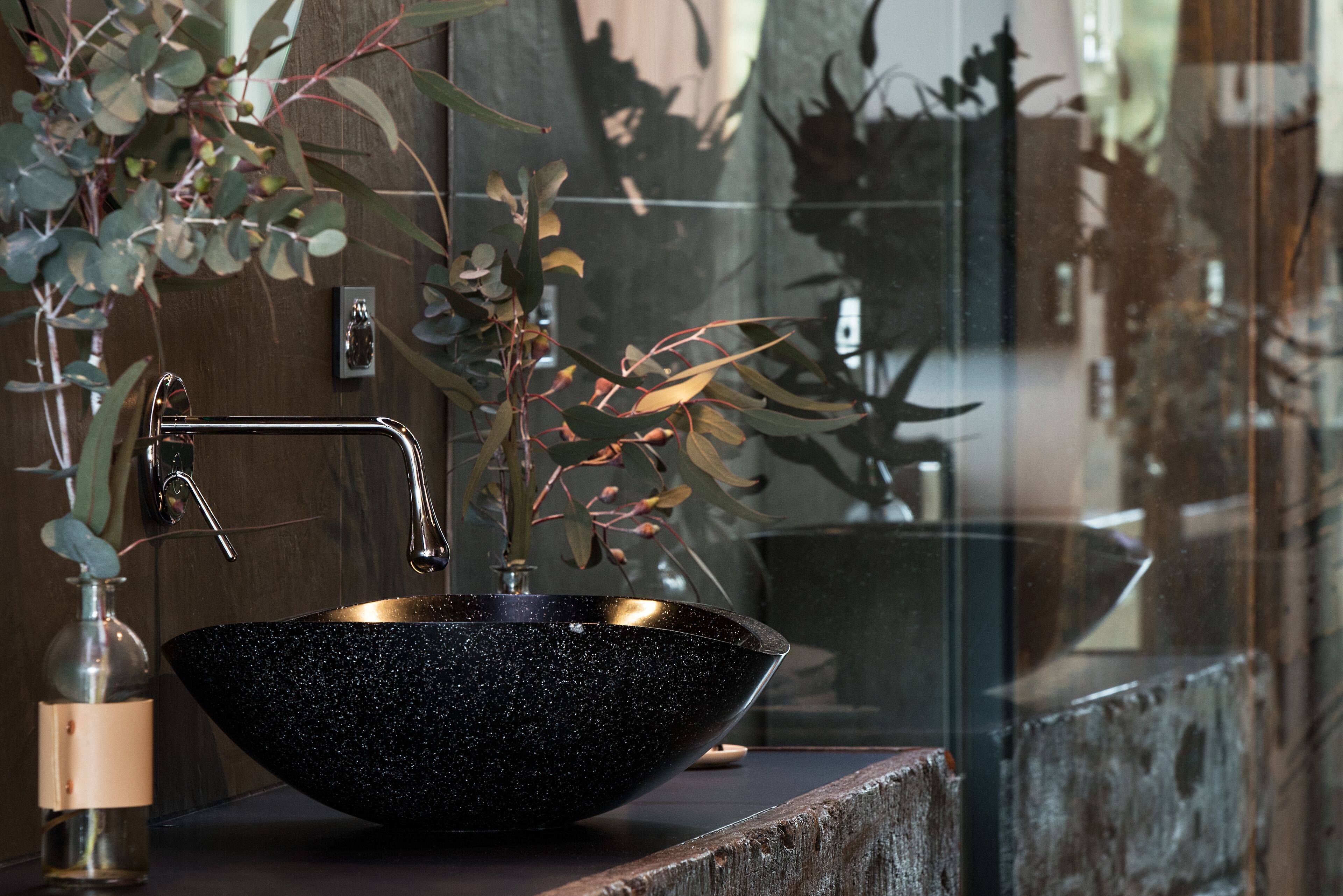 A black speckled bowl sink with a gold faucet, a glass vase with a plant, and a wooden ledge.