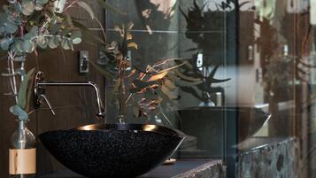 A black speckled bowl sink with a gold faucet, a glass vase with a plant, and a wooden ledge.