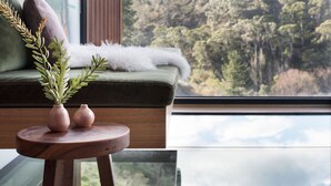 A cozy seating area with a green sofa, a small wooden table with two vases, and a view of a forest through a large window. - Ranked in the World's Top 60 Places to Elope (Hepburn Springs)