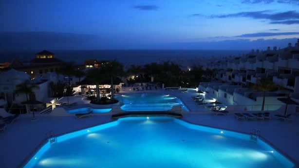 Una gran piscina iluminada con múltiples carriles, rodeada de sillas de descanso y palmeras, con vistas a la costa.