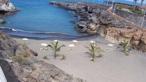 Una playa con terreno rocoso, sombrillas blancas y palmeras.