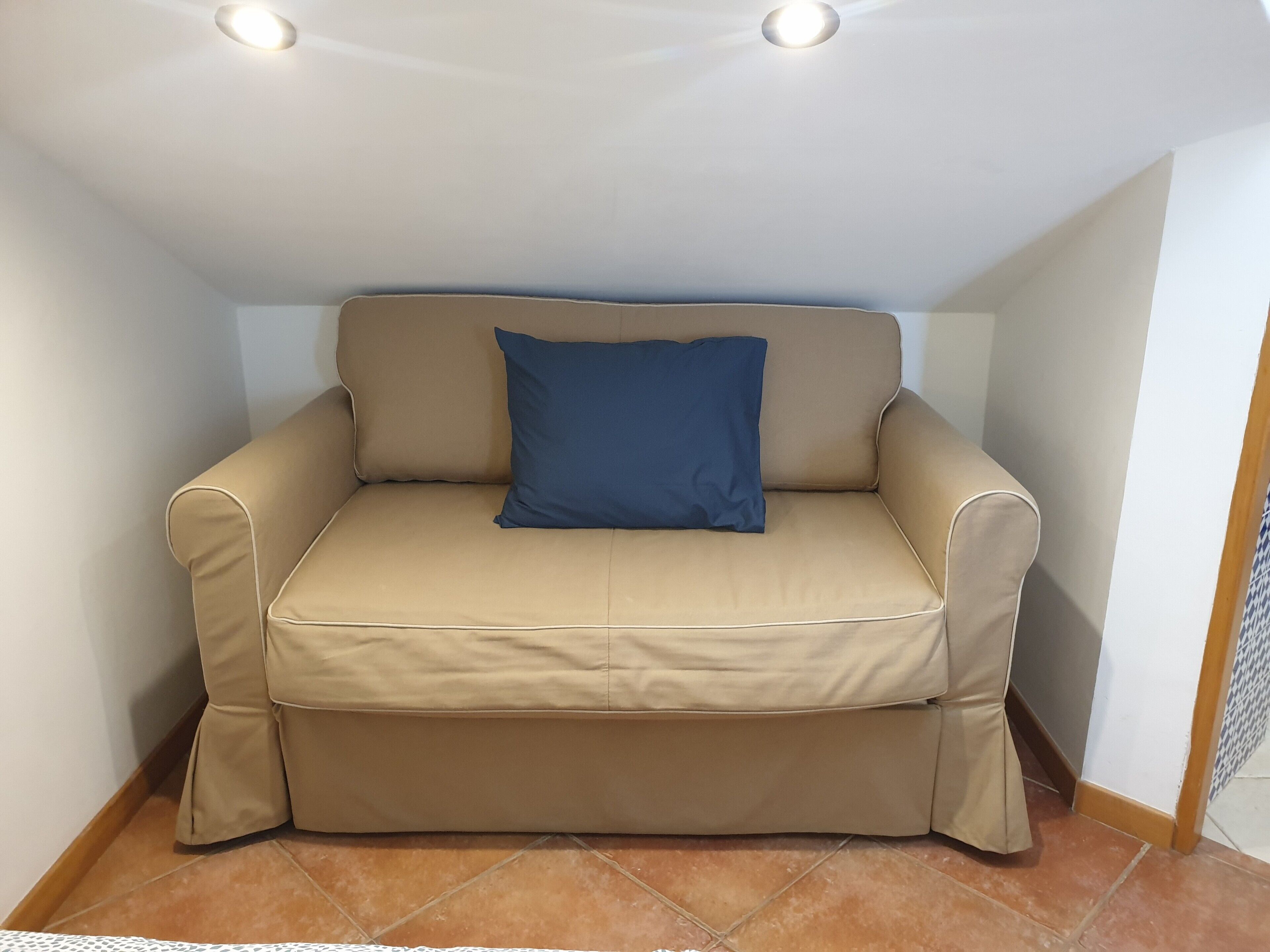 A beige sofa with a blue pillow in a corner of a room with white walls and a tiled floor.