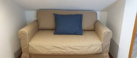 A beige sofa with a blue pillow in a corner of a room with white walls and a tiled floor.