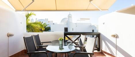 Ein Balkon mit rundem Tisch und Stühlen, einem Blumentopf und Blick auf weiße Gebäude mit Kuppeln.