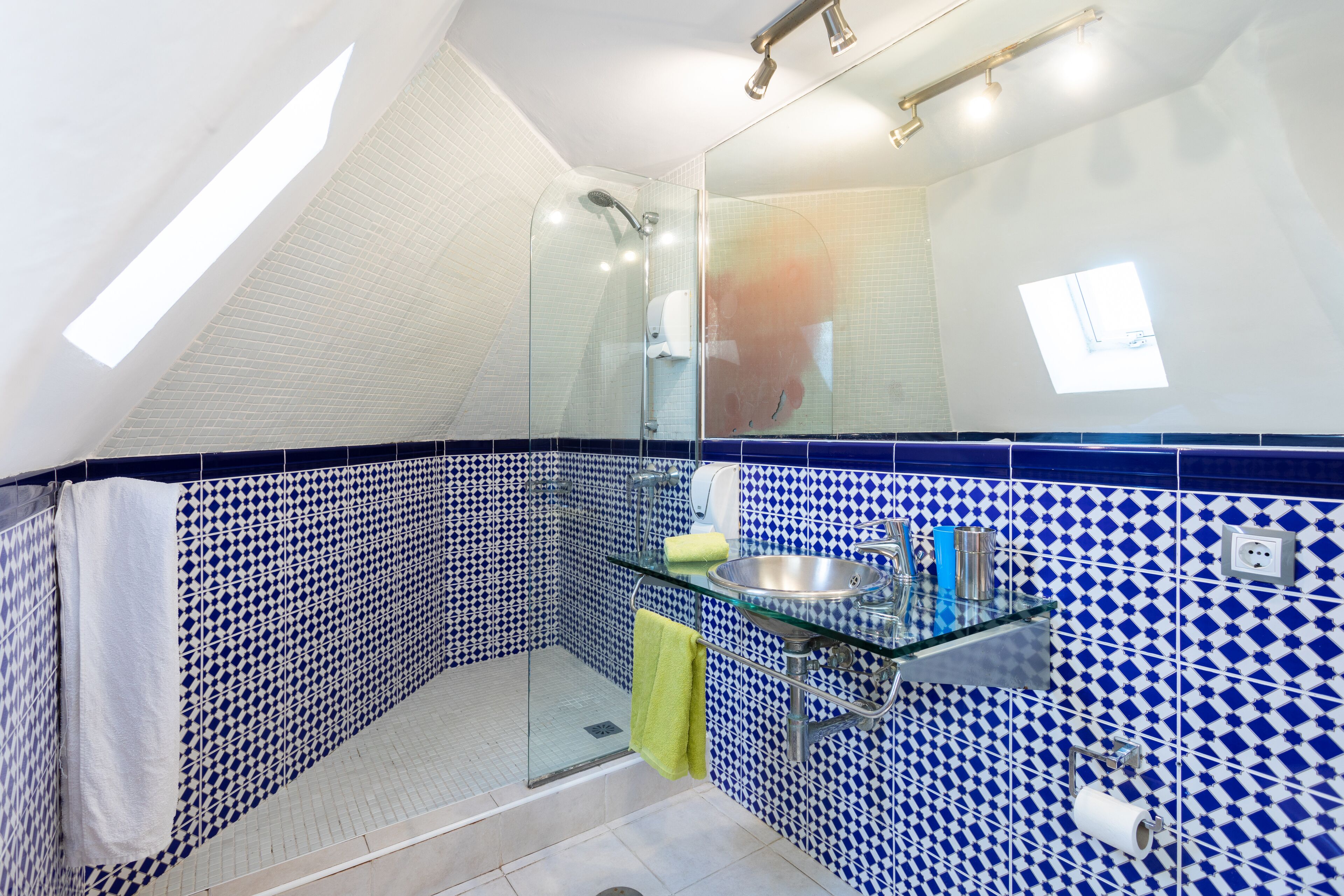 Casa de banho com chuveiro em cabine de vidro, lavatório de vidro e azulejos com padrão azul e branco.