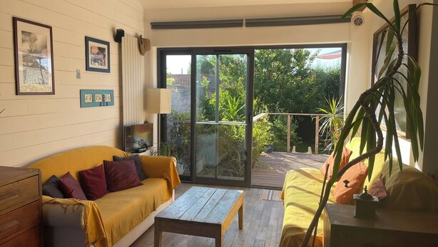 Ein Wohnzimmer mit einem gelben Sofa, einem Holztisch und einer Glastür, die auf einen Balkon mit Pflanzen und Blick auf Bäume führt.