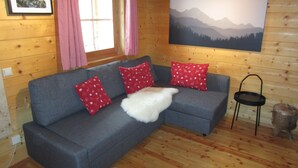 L’intérieur chaleureux d’un chalet avec un canapé gris, des coussins à motifs rouges, un plaid blanc et moelleux, un tableau encadré accroché au mur et une grande peinture de paysage.