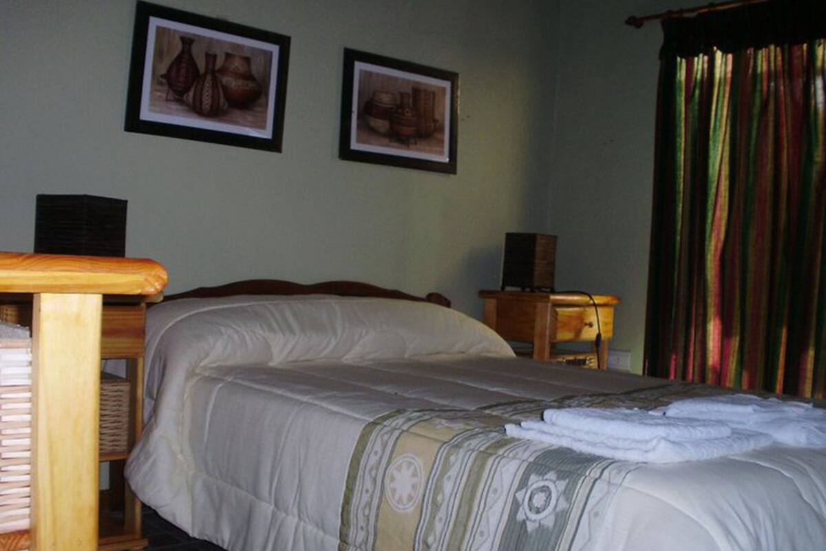 Una cama bien hecha con un edredón estampado, una mesita de noche con lámpara, y dos cuadros en la pared.