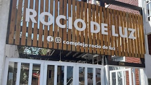 A wooden sign with "ROCIO DE LUZ" and social media icons, a white door with glass panels, and a building with a brick facade.