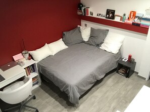 Ein ordentlich eingerichtetes Schlafzimmer mit einem Bett, einem Schreibtisch und einem Stuhl. Auf den Regalen befinden sich Bücher und persönliche Gegenstände.