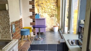 Eine schmale Küche mit einer Steinwand, einem violetten Tisch, einem gelben Hocker und einer Topfpflanze.