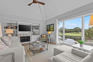 A modern living room with a fireplace, a flat-screen TV, a sofa, a coffee table, and a view of a golf course through large windows.