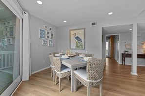 A dining area with a table set for four, surrounded by wicker chairs, a large framed artwork on the wall, and a view through a glass door.