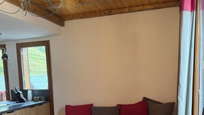 A cozy room with a wooden ceiling, a wooden bench with red and gray cushions, a wooden coffee table, and a window with curtains.