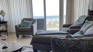 Un salon comprenant un canapé, des fauteuils et une table basse. De grandes fenêtres offrent une vue sur l’océan.