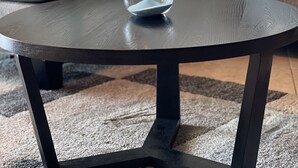 Une table ronde avec un sablier transparent placé au centre, un tapis à motifs et un canapé avec des coussins en arrière-plan.