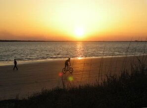 A beach at sunset with a person riding a bicycle and another walking.