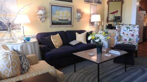 Una sala de estar con un sofá azul oscuro, un sillón estampado en amarillo y una mesa de centro con un jarrón de flores. Hay cuadros colgados en la pared y un espejo.