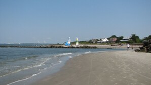 Una playa con un velero, un muelle a lo lejos y casas a lo largo de la costa.