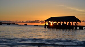 A pier with a covered structure at sunset, boats on the water, and a calm sea. - Kauai Sunrise, Rainbows, Ocean Views & 15th Fairway of Makai (Princeville)