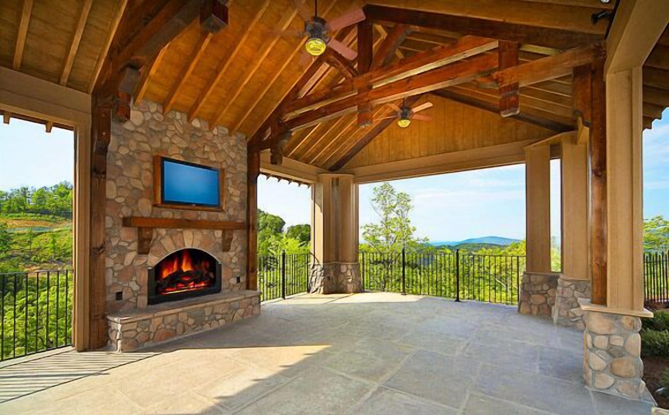 A covered outdoor patio with a stone fireplace, a television mounted on the wall, and a view of the surrounding landscape.
