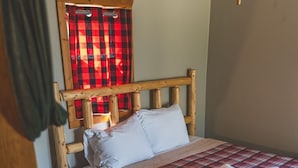 A bedroom with a wooden headboard, a plaid bedspread, two pillows, a bedside table, a window with red and black checkered curtains, and a ceiling fan. - Cozy 1BR Lakefront Cabin w/ Private Launch & Dock (Backus)