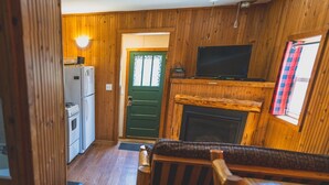 A cozy cabin interior with a wooden interior, a fireplace, a television, and a kitchenette. - Cozy 1BR Lakefront Cabin w/ Private Launch & Dock (Backus)