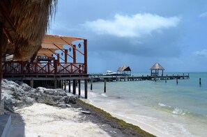 Une construction au bord de la plage, dotée d’un toit de chaume, une jetée en bois s’étendant dans l’eau et un bateau amarré à son extrémité.