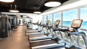 Ein Fitnessraum mit Laufbändern an einer Wand, große Fenster bieten Blick aufs Meer.