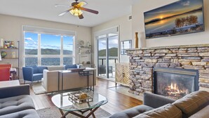 A spacious living room with a fireplace, a large window overlooking a body of water, and a ceiling fan.